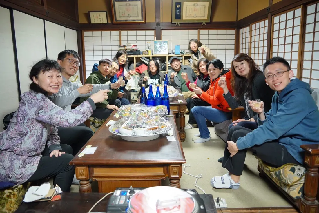 釀酒團的大家在酒造的和室裡圍著長桌舉杯,桌上有藍色酒瓶和料理
