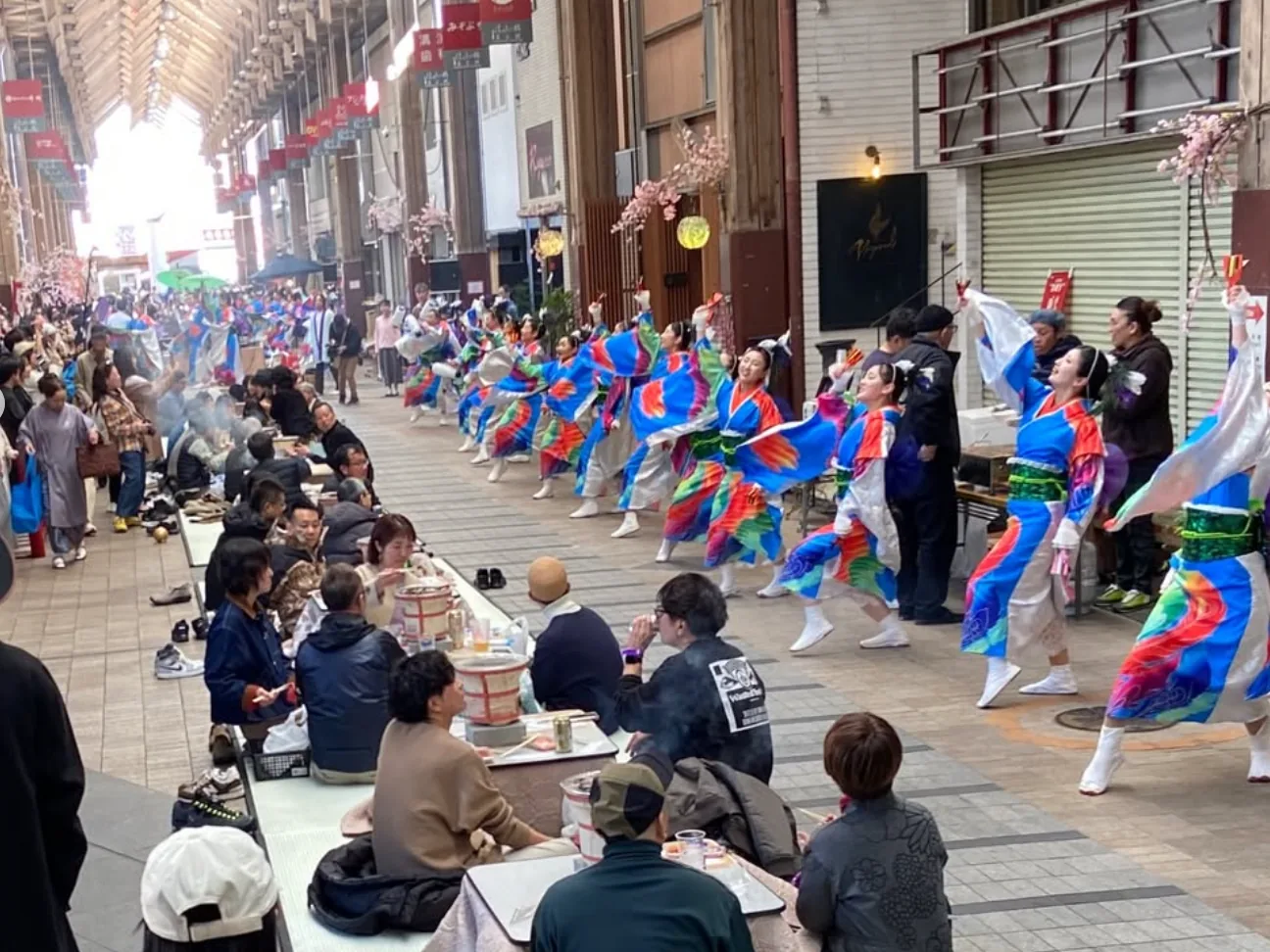 高知的商店街拱廊下,穿著彩色舞衣的よさこい舞者跳舞,旁邊的人坐在榻榻米上圍著小火爐吃喝